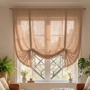 Op de afbeelding: Een gezellige woonkamer met een bruine leren bank, kussens en een geweven deken. Een groot raam is gedrapeerd met een beige linnen gordijn. Groene planten en een rieten mand voegen natuurlijke elementen toe.