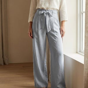 May include: A woman in a cream-coloured long-sleeved top and high-waisted, light blue trousers with a matching tie belt. The trousers have a wide-leg design. The outfit is made of a lightweight fabric. The woman is standing near a window.