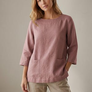 May include: A dusty rose-colored top with a wide, rounded neckline and three-quarter length sleeves. The garment features two square patch pockets on the front and a straight hemline, paired with light beige trousers in a neutral tone.
