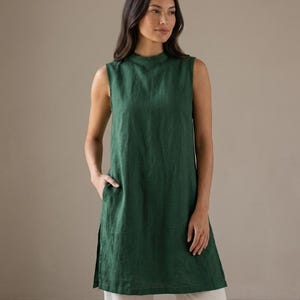 May include: A sleeveless forest green tunic featuring a high mock neckline, a straight silhouette, and side slits at the hem. The garment includes functional side pockets and is paired with light beige trousers, creating a simple, clean look.