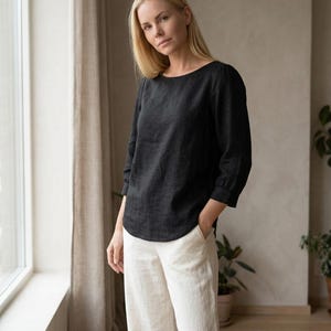 May include: A black long-sleeved top with a rounded neckline and a curved hemline, paired with off-white wide-leg trousers. The top features three-quarter length sleeves with cuffs, and the trousers have side pockets and a relaxed fit.