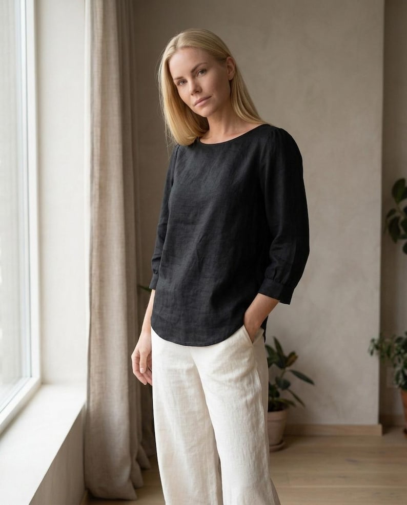 May include: A black long-sleeved top with a rounded neckline and a curved hemline, paired with off-white wide-leg trousers. The top features three-quarter length sleeves with cuffs, and the trousers have side pockets and a relaxed fit.