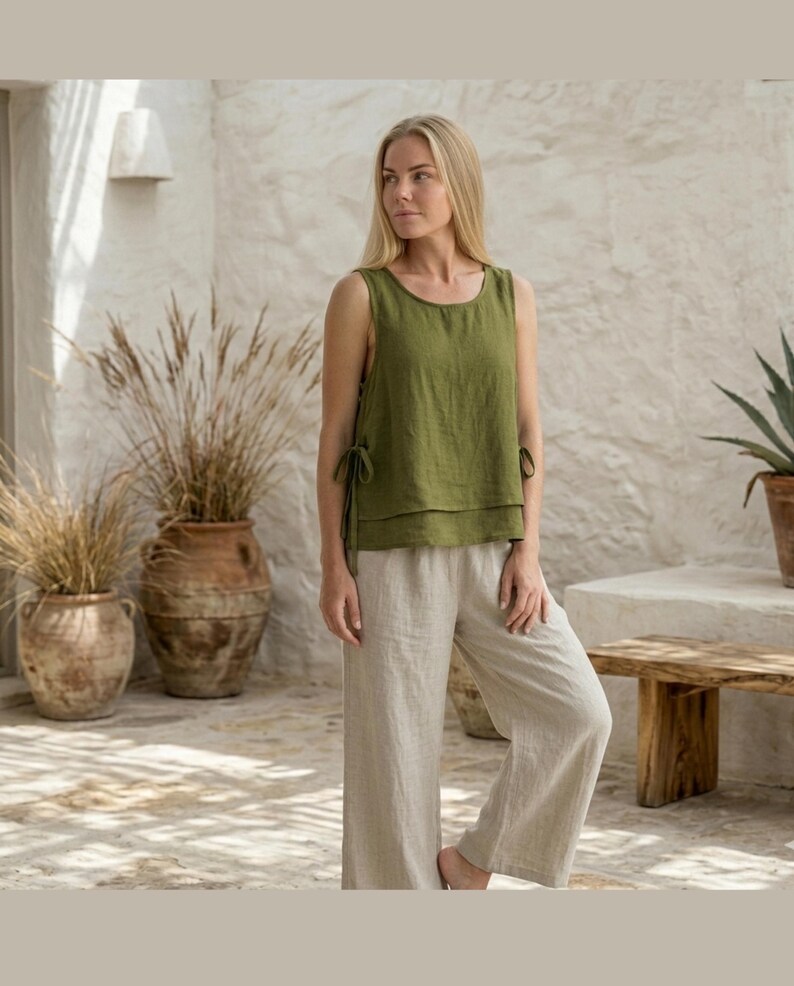 Puede incluir: Una mujer con un top sin mangas verde con detalles de lazo y pantalones de lino beige claro. El conjunto es informal y c&oacute;modo, perfecto para el clima c&aacute;lido. La parte superior es de color verde oliva liso y los pantalones son de color claro neutro.