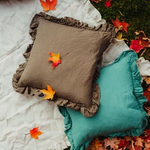 Puede incluir: Dos almohadas decorativas con bordes con volantes. Las almohadas son marrones y azul verdoso y est&aacute;n sobre una manta blanca con un patr&oacute;n a cuadros. La manta est&aacute; sobre un lecho de hojas de oto&ntilde;o.