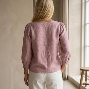 May include: A dusty rose long-sleeved blouse with a square neckline and gathered puff sleeves, paired with white trousers. The blouse features a straight hemline and buttoned cuffs, showcasing a textured fabric finish throughout the garment.