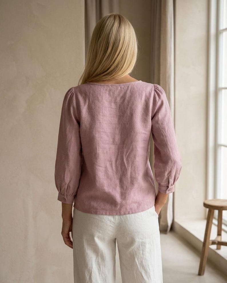 May include: A dusty rose long-sleeved blouse with a square neckline and gathered puff sleeves, paired with white trousers. The blouse features a straight hemline and buttoned cuffs, showcasing a textured fabric finish throughout the garment.