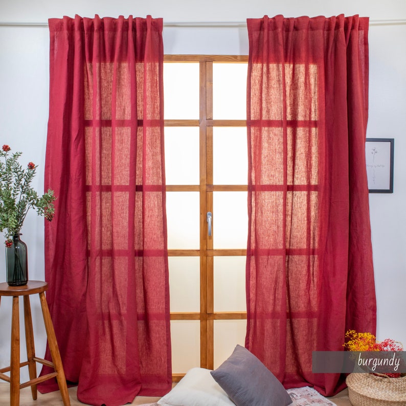 May include: Two burgundy linen curtains hanging in front of a window with a wooden frame. The curtains are sheer and have a subtle texture.