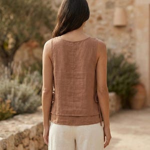 Puede incluir: Una blusa sin mangas de lino marr&oacute;n con un dise&ntilde;o en capas y lazos laterales. La modelo lleva la blusa con pantalones de lino color crema. El conjunto es adecuado para climas c&aacute;lidos y ocasiones informales.