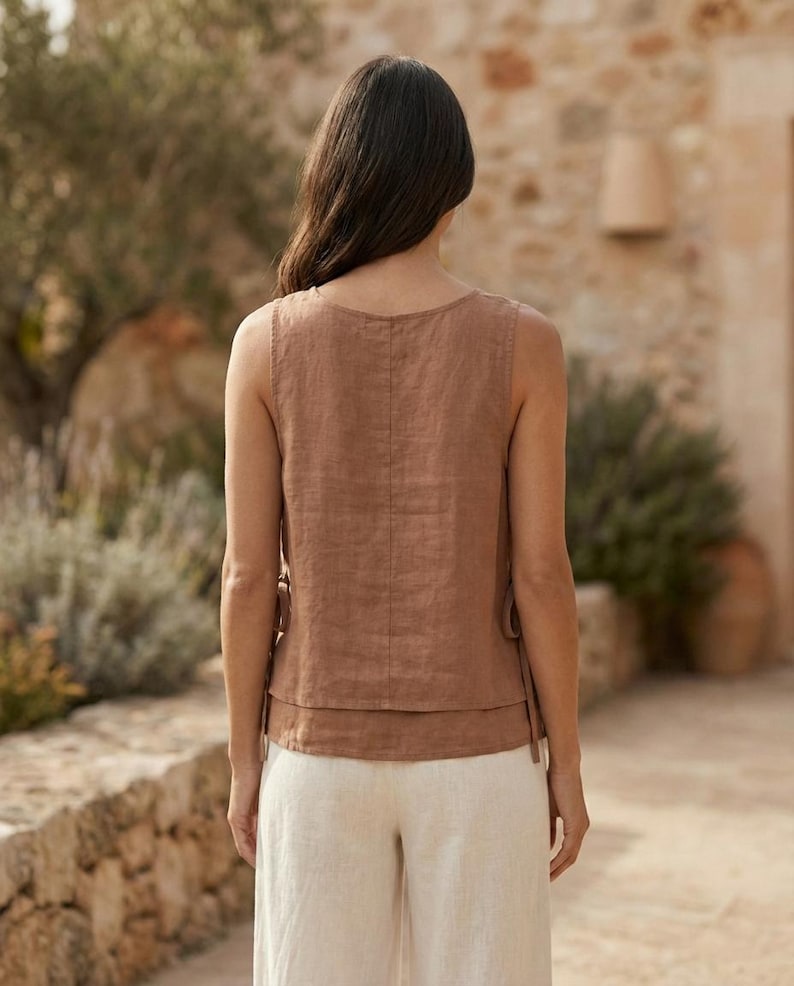 Puede incluir: Una blusa sin mangas de lino marr&oacute;n con un dise&ntilde;o en capas y lazos laterales. La modelo lleva la blusa con pantalones de lino color crema. El conjunto es adecuado para climas c&aacute;lidos y ocasiones informales.