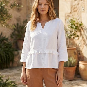 Könnte beinhalten: Ein weißes Leinenoberteil mit V-Ausschnitt und Dreiviertelärmeln. Das Oberteil hat eine Rüschendetail an der Taille und wird mit einer braunen Hose kombiniert. Das Outfit ist für warmes Wetter geeignet.