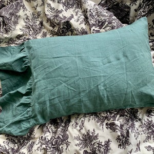 May include: A teal green pillow with a ruffled edge. The pillow is made of a soft, textured fabric and is lying on a white and black patterned fabric.