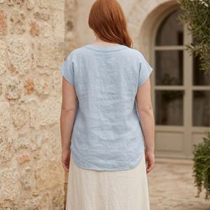 May include: A light blue short-sleeved top with a round neckline and a curved hemline, paired with a long, plain, off-white skirt. The top is made of a textured fabric and is shown from the back, displaying a simple, loose-fitting silhouette.