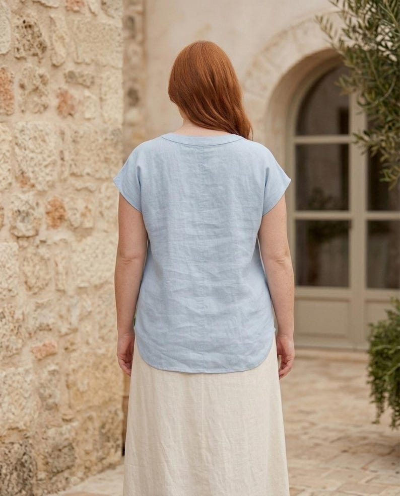 May include: A light blue short-sleeved top with a round neckline and a curved hemline, paired with a long, plain, off-white skirt. The top is made of a textured fabric and is shown from the back, displaying a simple, loose-fitting silhouette.