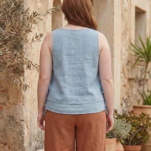Puede incluir: Una blusa sin mangas azul claro con un dise&ntilde;o en capas y detalles de lazo, combinada con pantalones de pierna ancha color &oacute;xido. El conjunto est&aacute; hecho de tela tipo lino, lo que sugiere un estilo casual y c&oacute;modo. El modelo est&aacute; al aire libre.