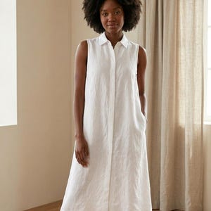 May include: A sleeveless white midi dress featuring a classic collar, a full-length button-down front placket, and side pockets. The garment is constructed from a textured fabric and falls to a straight hemline, maintaining a clean and simple silhouette.