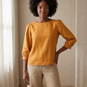 May include: A mustard yellow long-sleeved blouse with a boat neckline and gathered puff sleeves, paired with light beige wide-leg trousers. The blouse features a straight hemline and cuffed sleeves, while the trousers have a simple waistband.
