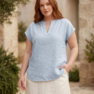 May include: A light blue short-sleeved blouse featuring a notched V-neckline and a relaxed fit. The garment is crafted from a textured fabric and includes a curved hemline, paired with a plain off-white skirt that reaches below the knees.