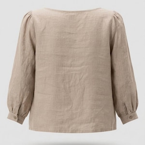 May include: A beige long-sleeved blouse with a wide neckline and gathered shoulders. The sleeves feature cuffs with a single button closure. The garment is made of a textured fabric and has a straight hemline, providing a simple and clean look.