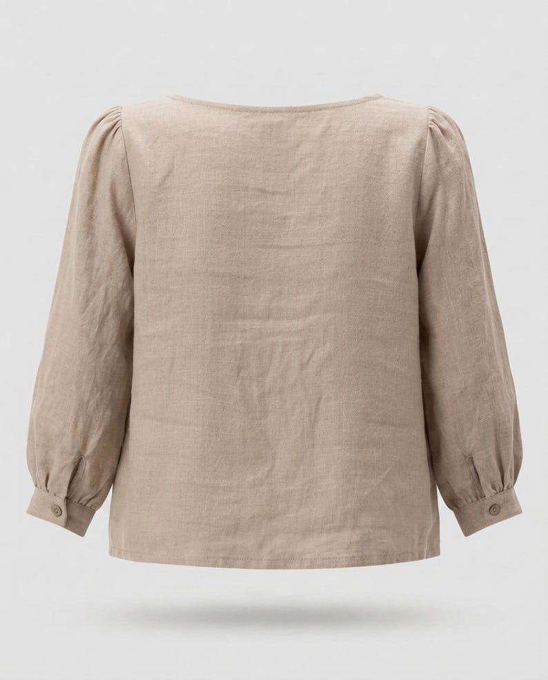 May include: A beige long-sleeved blouse with a wide neckline and gathered shoulders. The sleeves feature cuffs with a single button closure. The garment is made of a textured fabric and has a straight hemline, providing a simple and clean look.