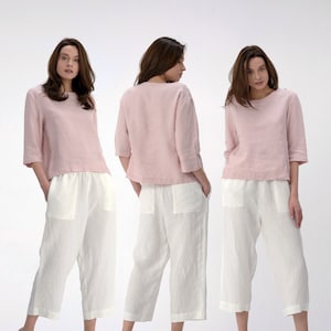 Pu&ograve; includere: Una vista a tre quarti di una donna che indossa una blusa di lino rosa e pantaloni di lino bianchi. La blusa ha una vestibilit&agrave; rilassata e una scollatura rotonda. I pantaloni hanno una gamba larga e una lunghezza a met&agrave; polpaccio.