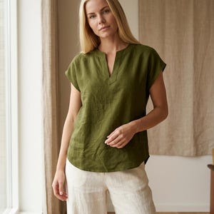 May include: An olive green short-sleeved blouse featuring a notched neckline and a relaxed fit. The top is constructed from a textured fabric and is paired with light beige trousers that have a straight-leg cut and a clean, simple design.