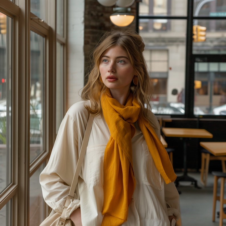 May include: A woman wearing a white shirt with a mustard yellow scarf tied around her neck. She is standing in front of a window and looking to the left.