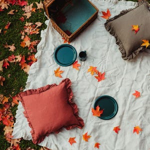 Puede incluir: Una manta de picnic blanca con una cesta de mimbre, dos almohadas, dos platos y una taza. La manta est&aacute; rodeada de hojas de oto&ntilde;o ca&iacute;das.