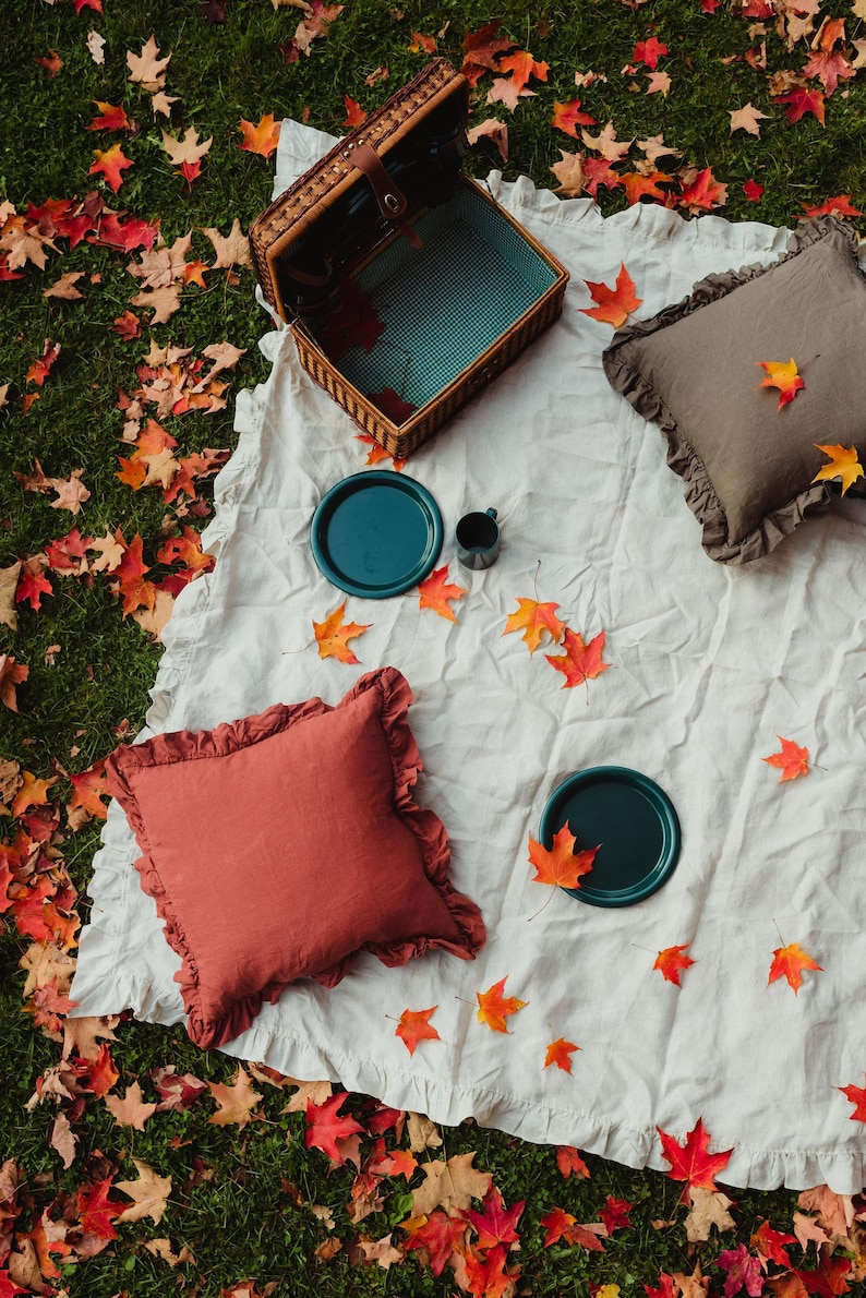 Puede incluir: Una manta de picnic blanca con una cesta de mimbre, dos almohadas, dos platos y una taza. La manta est&aacute; rodeada de hojas de oto&ntilde;o ca&iacute;das.