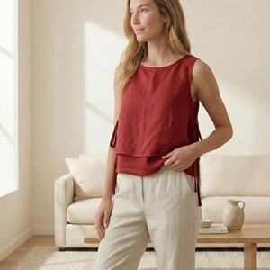 Puede incluir: Un top sin mangas de color rojo ladrillo con lazos laterales y un dise&ntilde;o en capas. Combinado con pantalones de lino beige claro. El atuendo se usa en una habitaci&oacute;n bien iluminada con una paleta de colores neutros.