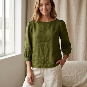 May include: An olive green long-sleeved blouse with a boat neckline and gathered puff sleeves at the shoulders, paired with off-white trousers. The blouse features a straight hemline and a textured fabric finish, creating a simple and clean look.