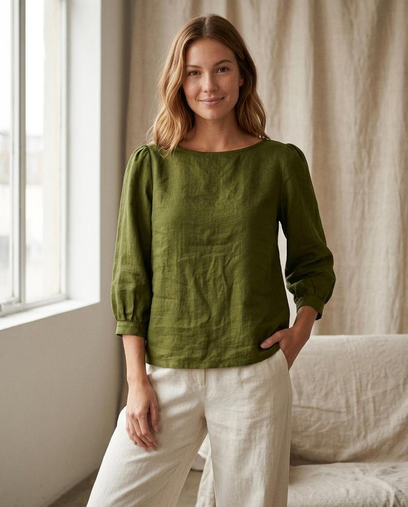 May include: An olive green long-sleeved blouse with a boat neckline and gathered puff sleeves at the shoulders, paired with off-white trousers. The blouse features a straight hemline and a textured fabric finish, creating a simple and clean look.