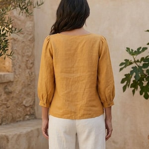 May include: A mustard yellow blouse with three-quarter length puff sleeves and buttoned cuffs, paired with off-white trousers. The blouse features a wide neckline and a straight hem, showcasing a textured fabric finish throughout the garment.