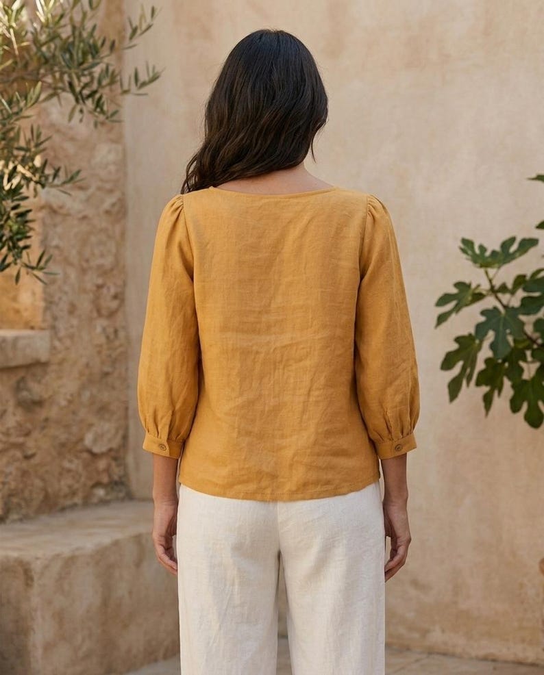 May include: A mustard yellow blouse with three-quarter length puff sleeves and buttoned cuffs, paired with off-white trousers. The blouse features a wide neckline and a straight hem, showcasing a textured fabric finish throughout the garment.