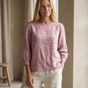 May include: A dusty rose-colored long-sleeve blouse featuring a boat neckline, slightly puffed shoulders, and gathered cuffs at the three-quarter length sleeves. The garment is paired with off-white trousers, creating a neutral-toned ensemble.