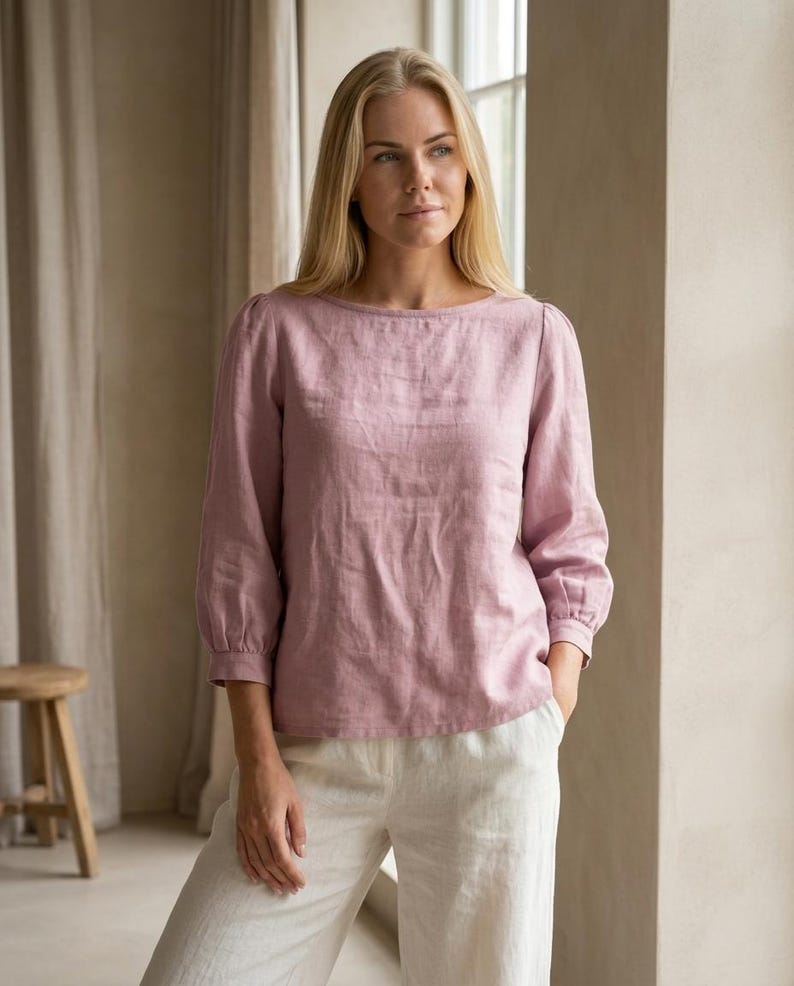 May include: A dusty rose-colored long-sleeve blouse featuring a boat neckline, slightly puffed shoulders, and gathered cuffs at the three-quarter length sleeves. The garment is paired with off-white trousers, creating a neutral-toned ensemble.