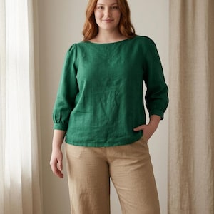 May include: A long-sleeved emerald green blouse with a boat neckline and gathered sleeve cuffs, paired with tan trousers. The blouse features a relaxed fit and a textured fabric appearance, while the trousers have a straight-leg silhouette.