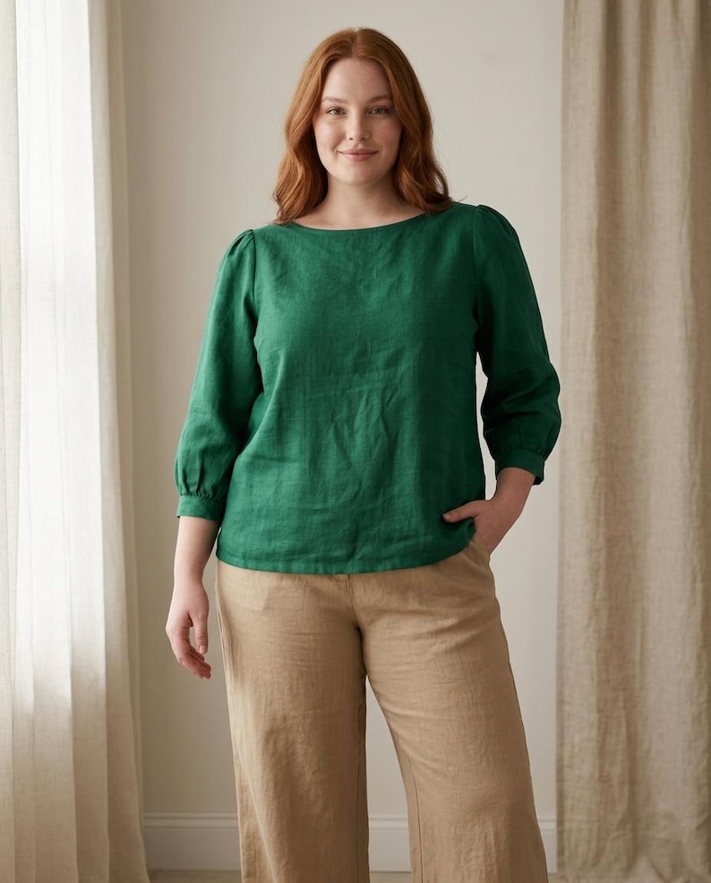 May include: A long-sleeved emerald green blouse with a boat neckline and gathered sleeve cuffs, paired with tan trousers. The blouse features a relaxed fit and a textured fabric appearance, while the trousers have a straight-leg silhouette.