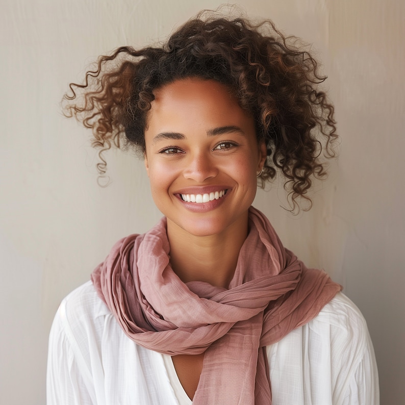 May include: A woman with curly brown hair smiles while wearing a white shirt and a pink scarf.