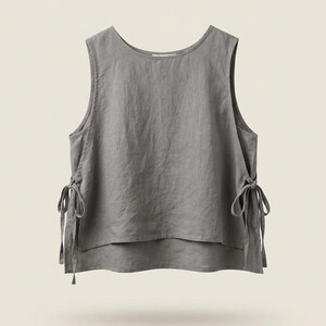 Puede incluir: Una blusa sin mangas de lino gris con cuello redondo y lazos laterales. La blusa tiene un dise&ntilde;o en capas y un ajuste relajado. La tela parece suave y ligeramente texturizada.