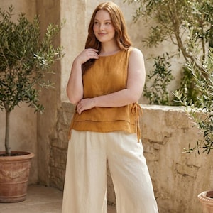 Puede incluir: Top sin mangas amarillo mostaza con dise&ntilde;o en capas y lazos laterales, combinado con pantalones de pierna ancha color crema. El conjunto est&aacute; hecho de una tela ligera, perfecto para el clima c&aacute;lido. El modelo est&aacute; de pie frente a una pared de piedra y olivos.