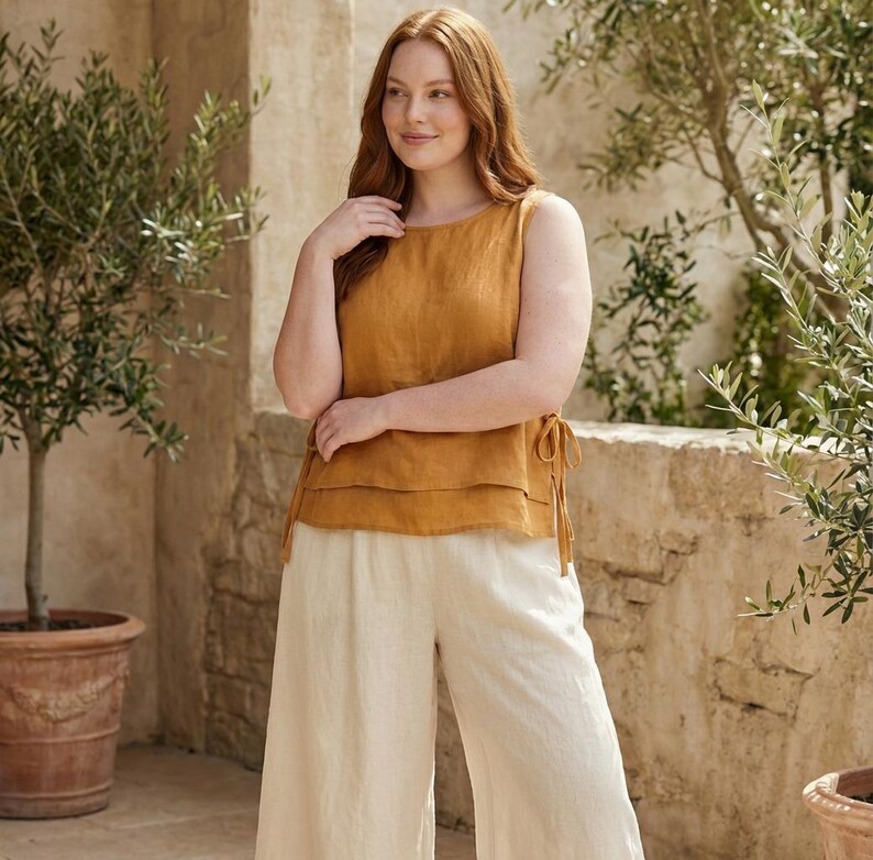 Puede incluir: Top sin mangas amarillo mostaza con dise&ntilde;o en capas y lazos laterales, combinado con pantalones de pierna ancha color crema. El conjunto est&aacute; hecho de una tela ligera, perfecto para el clima c&aacute;lido. El modelo est&aacute; de pie frente a una pared de piedra y olivos.