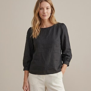 May include: A charcoal gray long-sleeved blouse featuring a round neckline and gathered sleeve cuffs. The top is paired with light beige trousers. The blouse has a straight hemline and a simple, clean design with a textured fabric appearance.