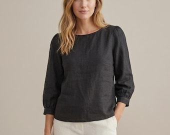 May include: A charcoal gray long-sleeved blouse featuring a round neckline and gathered sleeve cuffs. The top is paired with light beige trousers. The blouse has a straight hemline and a simple, clean design with a textured fabric appearance.