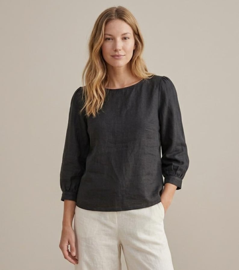 May include: A charcoal gray long-sleeved blouse featuring a round neckline and gathered sleeve cuffs. The top is paired with light beige trousers. The blouse has a straight hemline and a simple, clean design with a textured fabric appearance.