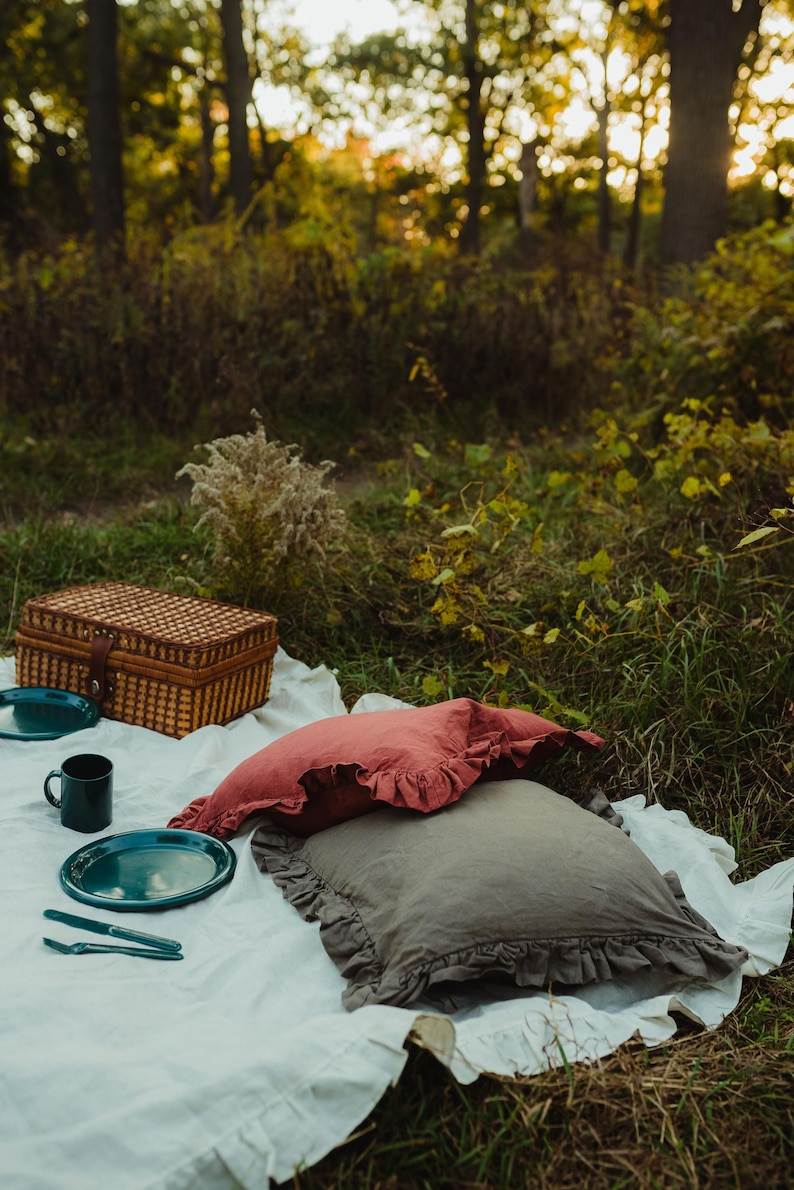 Puede incluir: Una manta de picnic con una cesta de mimbre, dos almohadas, dos platos verdes y una taza verde. La manta est&aacute; extendida en el suelo en un entorno boscoso.