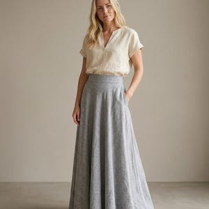 May include: A beige short-sleeved blouse with a split neckline and cuffed sleeves paired with a high-waisted, floor-length grey maxi skirt. The skirt features a wide waistband, a flared A-line silhouette, and side pockets, creating a simple ensemble.