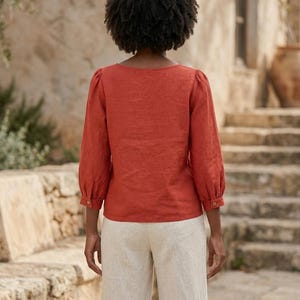 May include: A rust-colored long-sleeve blouse with a square neckline and gathered shoulders, paired with light beige trousers. The blouse features three-quarter length sleeves with buttoned cuffs, providing a structured yet simple silhouette.