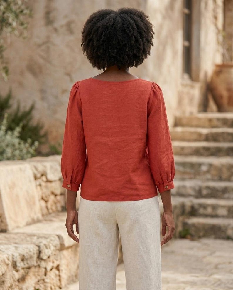 May include: A rust-colored long-sleeve blouse with a square neckline and gathered shoulders, paired with light beige trousers. The blouse features three-quarter length sleeves with buttoned cuffs, providing a structured yet simple silhouette.