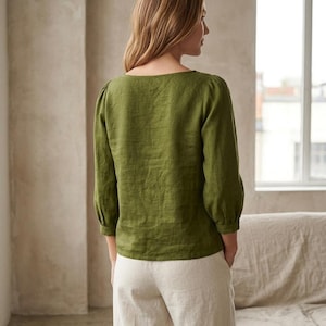 May include: An olive green long-sleeved blouse with a square neckline and gathered sleeve cuffs, paired with light beige trousers. The blouse features a straight hemline and a textured fabric finish, worn with the hands tucked into the trouser pockets.