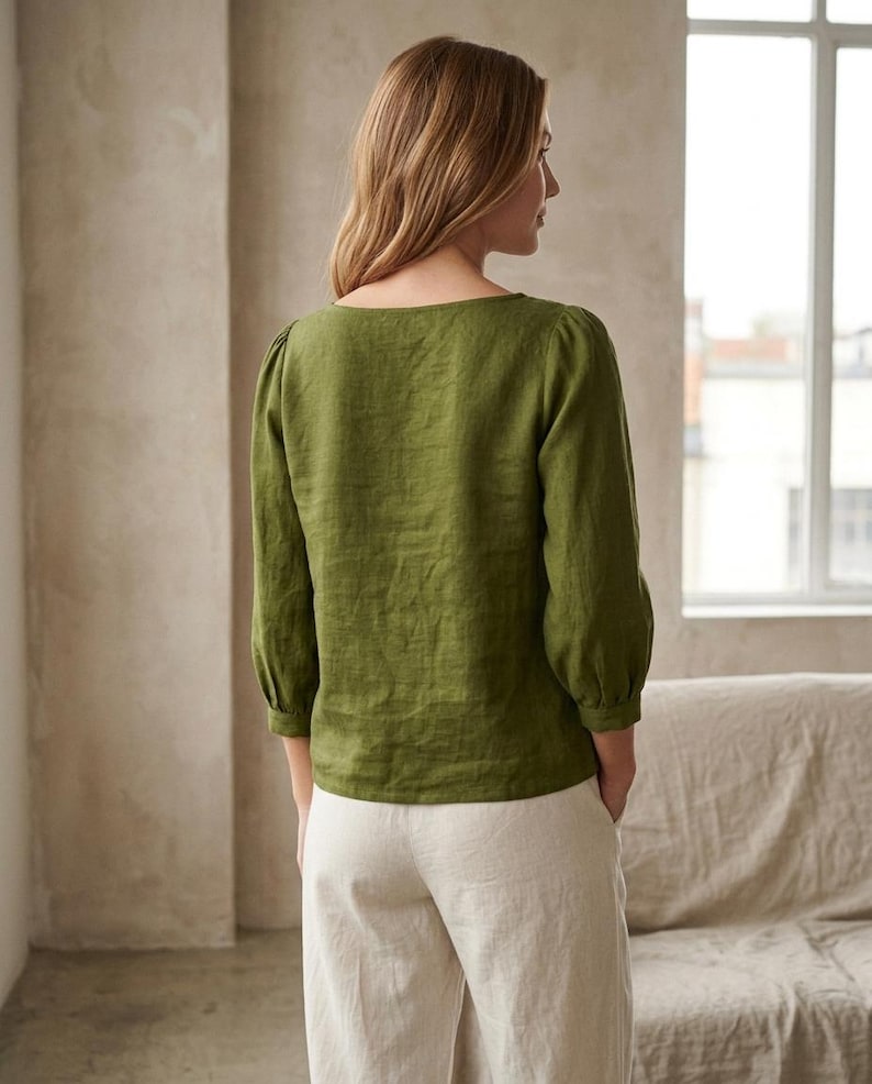 May include: An olive green long-sleeved blouse with a square neckline and gathered sleeve cuffs, paired with light beige trousers. The blouse features a straight hemline and a textured fabric finish, worn with the hands tucked into the trouser pockets.